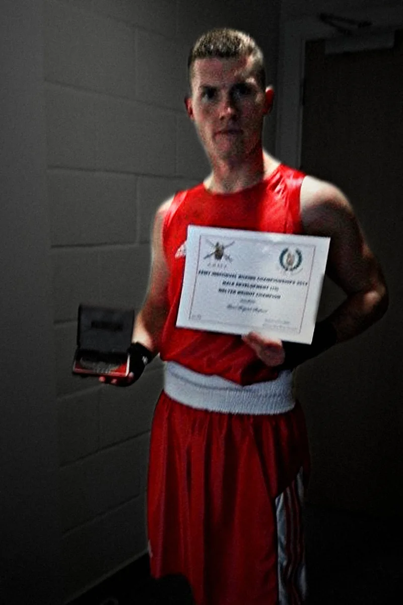 A boxer in Army red holding his medal and the 2014 Army Individual Boxing Championships certificate